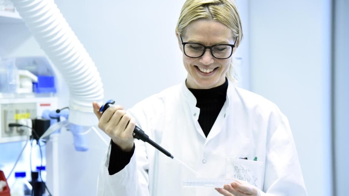 Miriam står i hvid kittel i et laboratorium med en pipette i hånden 