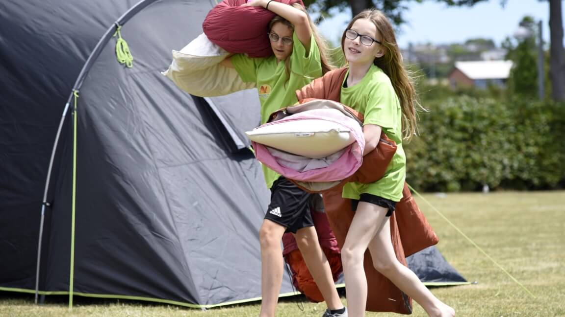 To piger kommer slæbende med sovepose og puder. Den ene bærer på hovedet og den anden har favnet fyldt. De har begge grønne T-shirts på fra sommerskolen