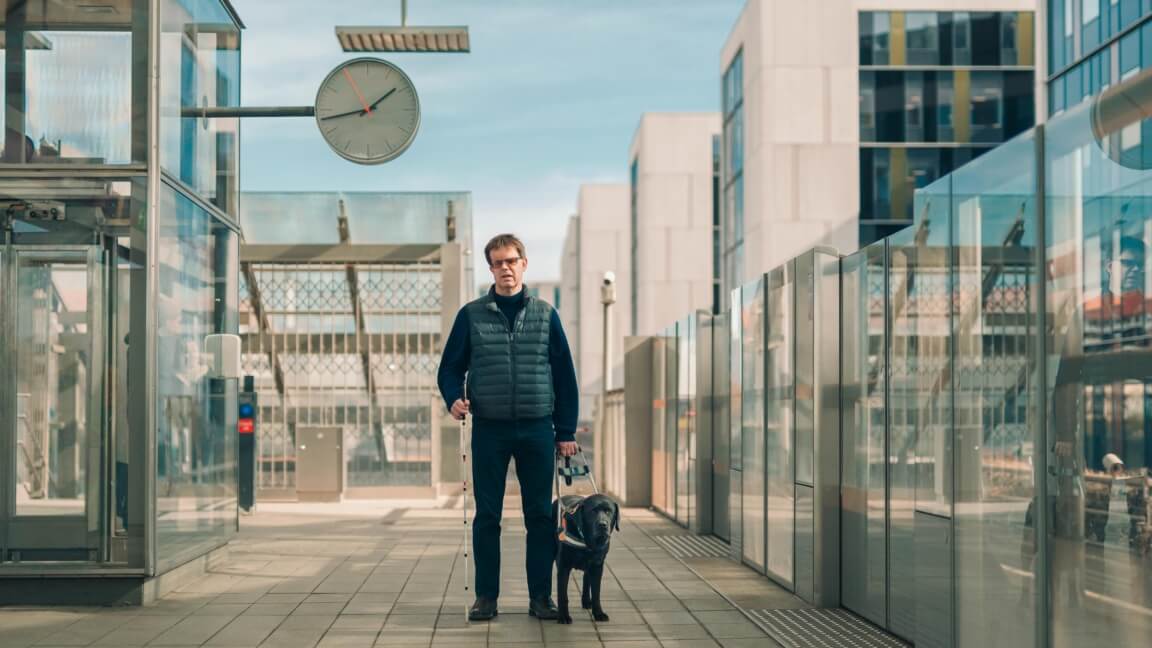 Bue står på en metrostation med en blindestok i den ene hånden og førerhunden Buller, en sort labrador, i den anden hånd