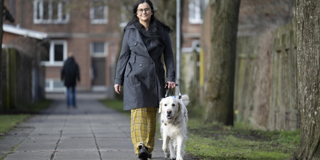 Tijana går på fortovet med førerhunden Buster, en lys golden retriever iført førerhundebøjle