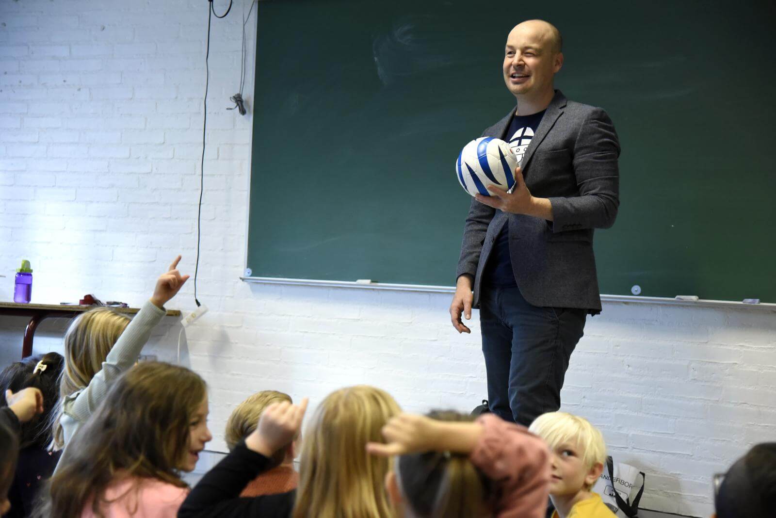 Ask holder en fodbold foran en skoleklasse, der sidder på gulvet