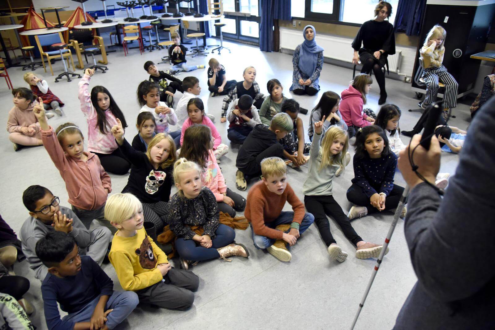 En masse skoleelever sidder på gulvet, tre af børnene sidder med fingeren oppe