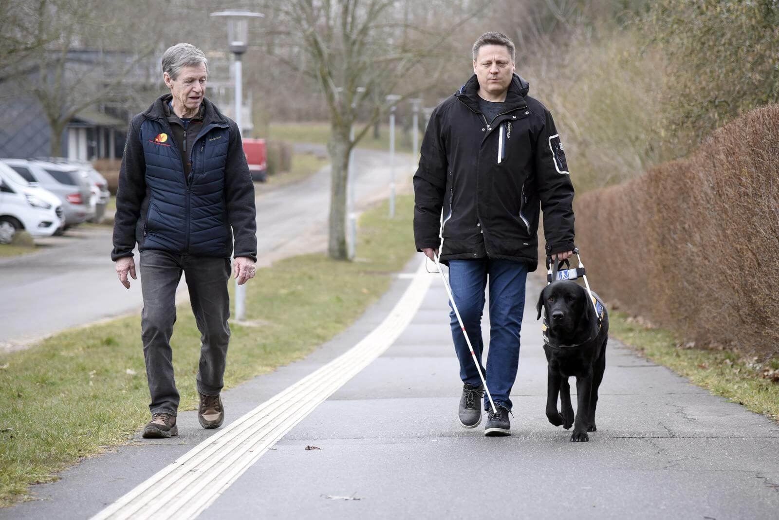 Kenneth går med en sort labrador i førerhundebøjle og ved siden af går en mandlig førerhundeinstruktør