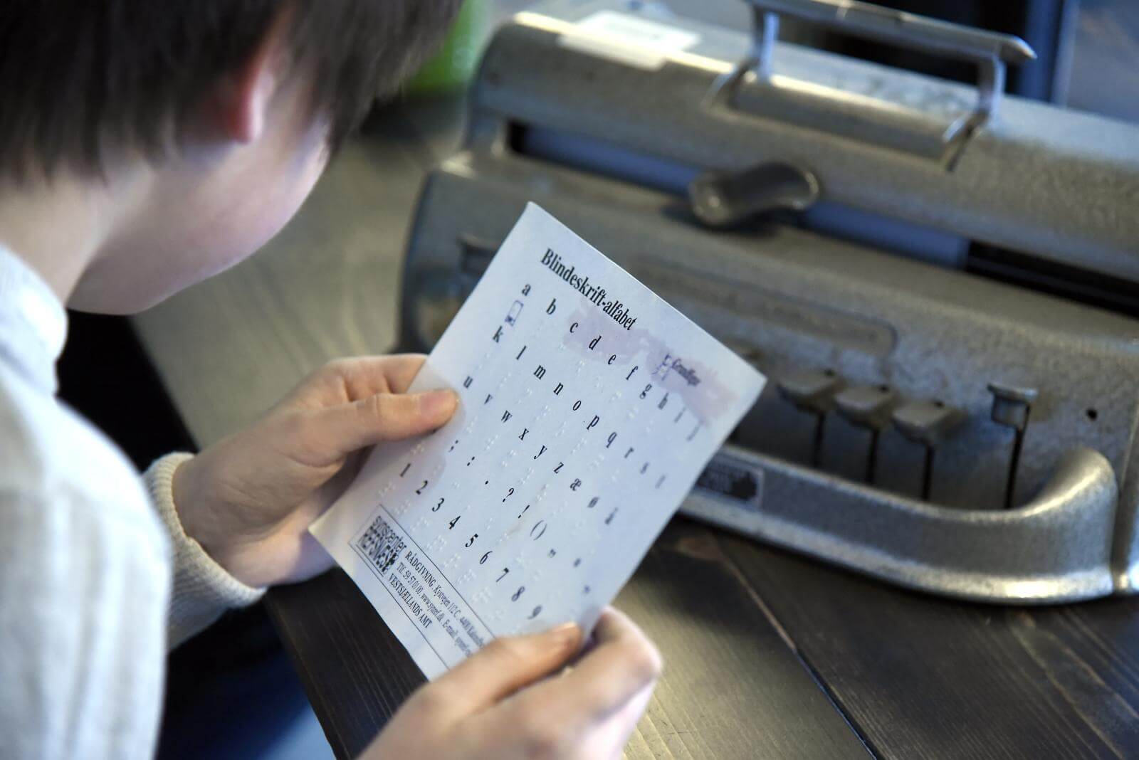 Gustav sidder med et ark med Braille alfabetet og en Perkins punktskriftmaskine i baggrunden 