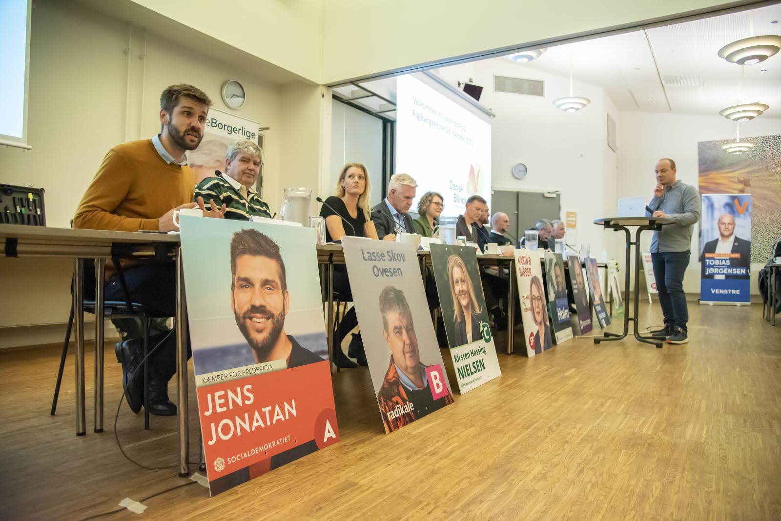 Folketingskandidaterne sidder ved et langbord og kigger ud mod tilhørerne. Landsformand Ask Løvbjerg Abildgaard står ved et højt cafebord mellem kandidaterne og tilhørerne. 