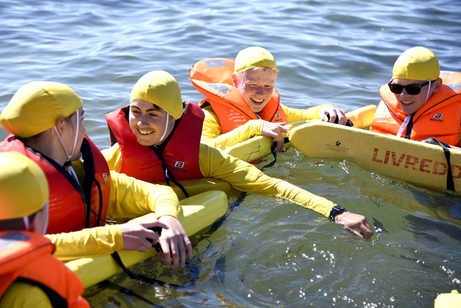 Fem børn flyder i vandet med stort smil og på et gult bræt, hvor der står livredder. De er alle iført våddragter, redningsveste og gule badehætter