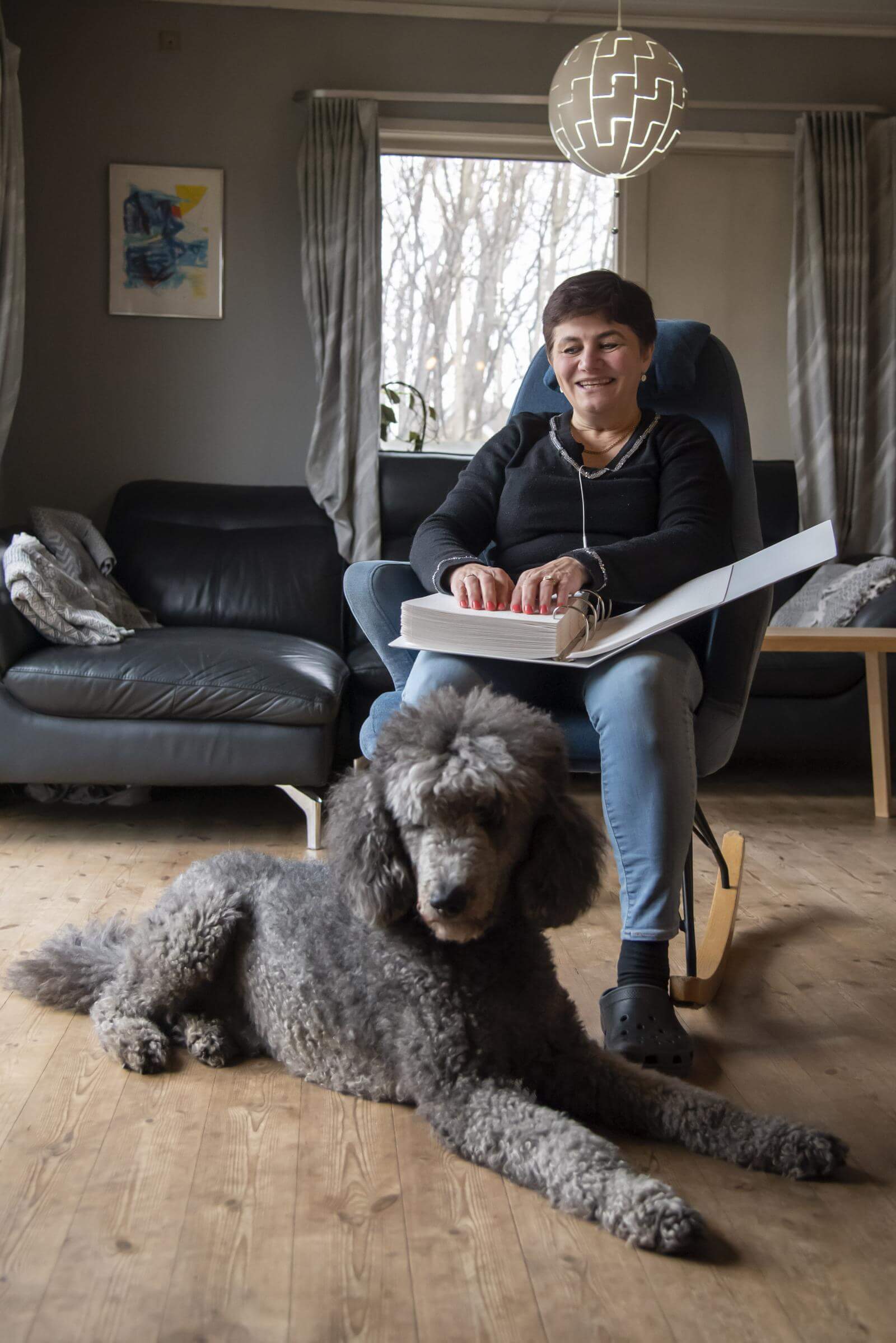 Jannie læser punktskrift i in gyngestol med førerhunde Kingo, en grå storpuddel, liggende for fødderne