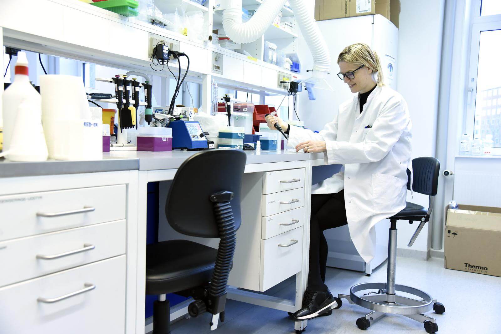 Miriam sidder i hvid kittel i et laboratorium med en pipette i hånden 