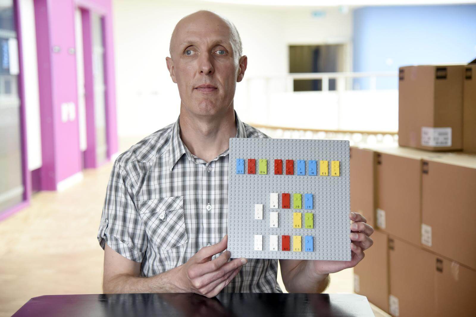 Christian viser en plade med Lego Braille Bricks, hvor der står god sommer. I baggrunden anes en masse kasser med Lego Braille Bricks