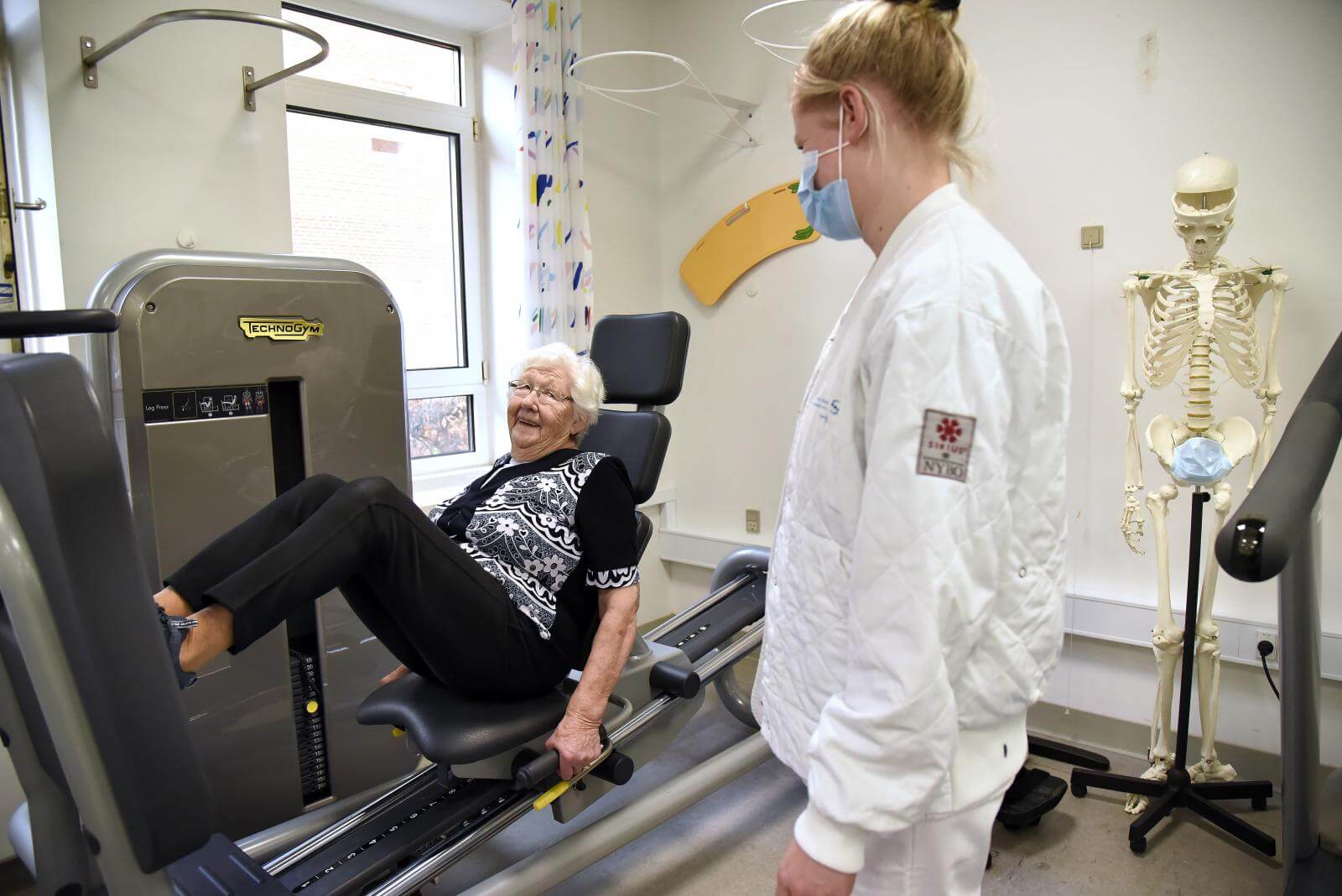 88-årige Gurli løfter kilo i benpres, mens hun smilende kigger på fysioterapeuten 