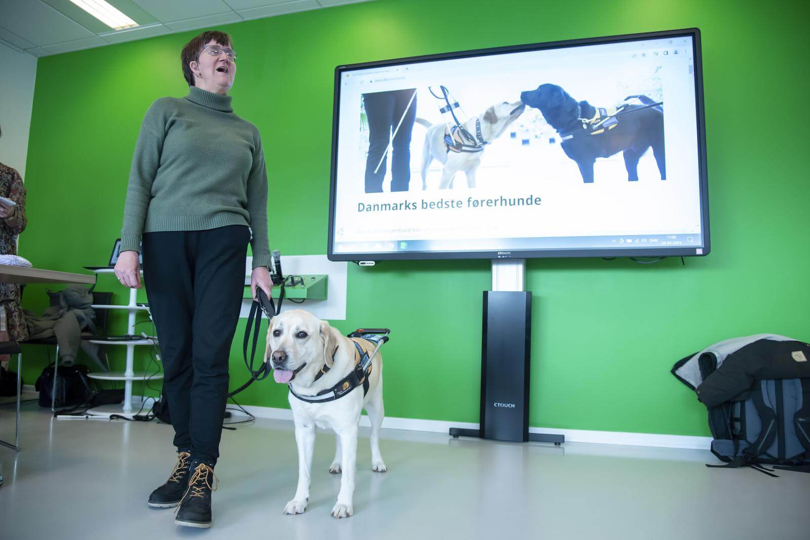Lene og førerhunden Ronja står foran en skærm, der viser billede af førerhunde 