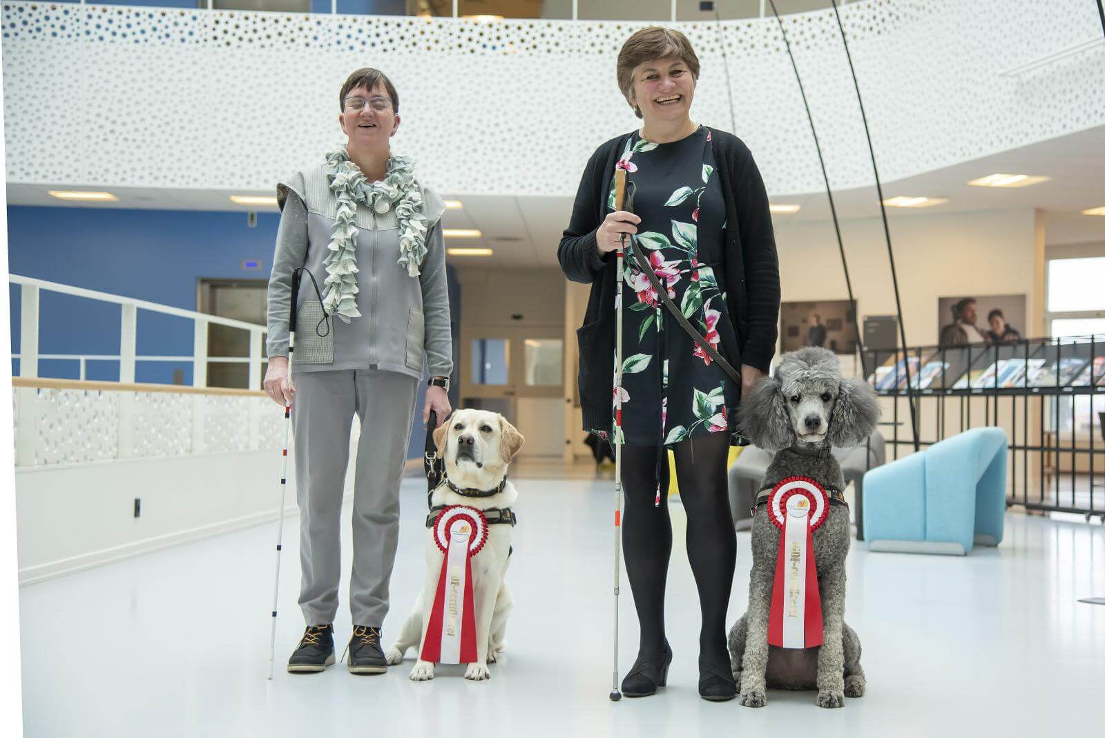 Jannie og Lene står ved siden af hinanden med en blindestok i hånden. Ved siden af dem sidder førerhunden Ronja, lys labrador og førerhunden Kingo, grå storpuddel, med en stor roset på brystet hver