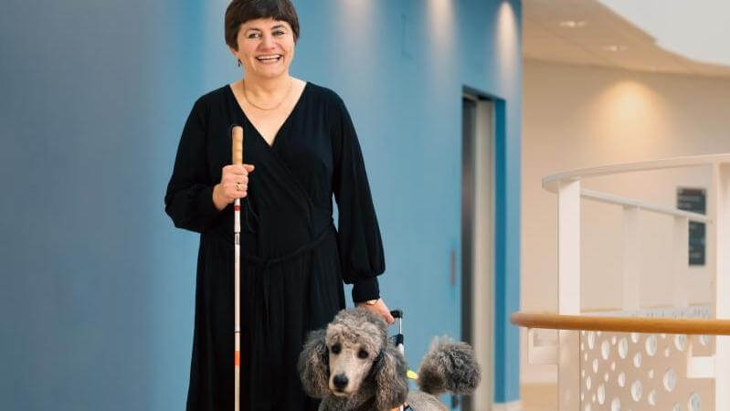 Kvinde med blindestok står på indenfor sammen med en førerhund foran en blå væg
