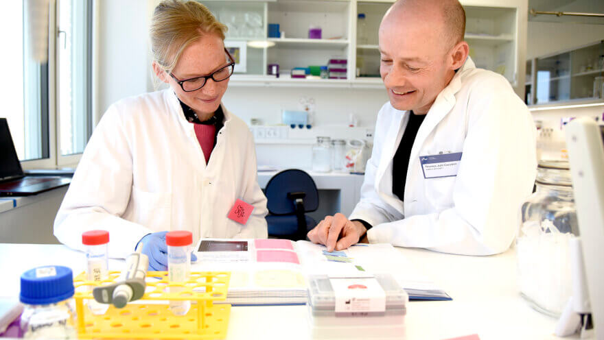 Silja og Thomas Corydon sidder i laboratoriet iført hvide kitler og studerer i en fagbog