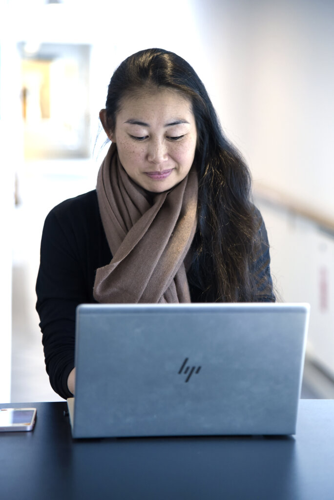 En ung dame sidder foran sin computer og arbejder. Ved siden af computeren ligger en iphone. Hun ser koncentreret ud.