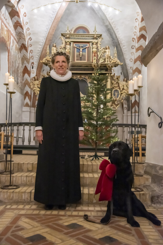 Rita står foran alteret iført præstekjole. Ved siden af sidder hendes førerhund, en sort labrador med en stor rødjulesløjfe om halsen. I baggrunden ses et juletræ
