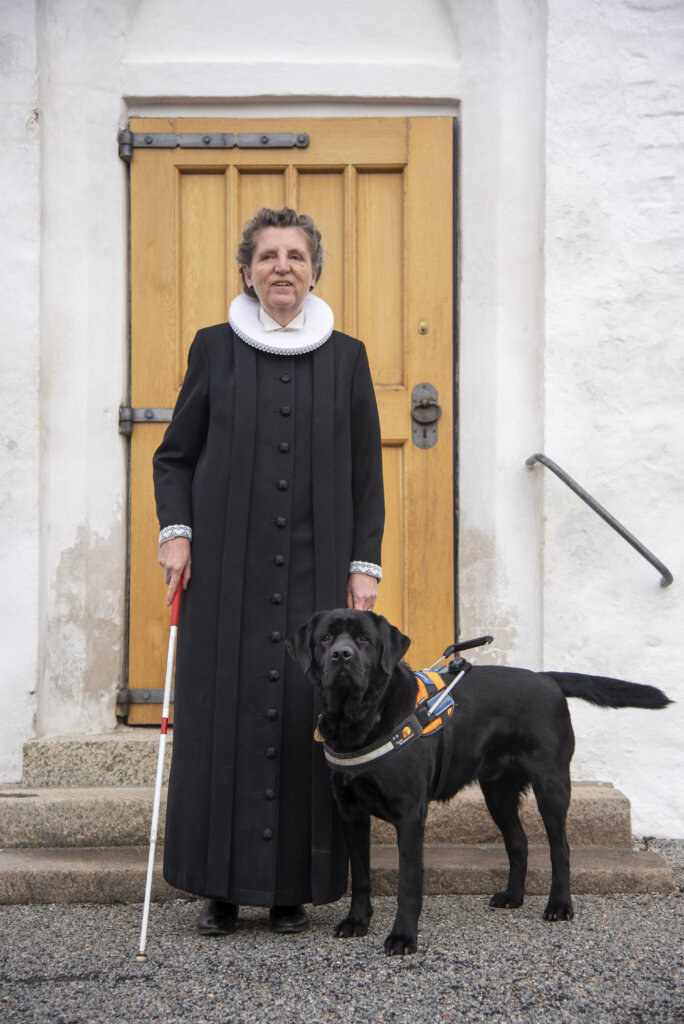 Rita er iført præstekjole og blindestok i den ene hånd. Ved siden af står hendes førerhund, en sort labrador iført førerhundebøjle. De stå ude foran kirkedøren