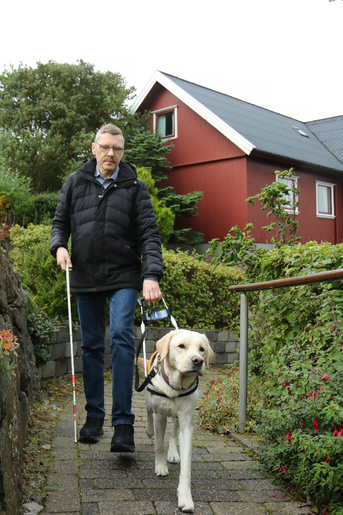 Johan går med førerhunden Milo, en lys labrador, op ad en havesti. I baggrunden ses et rødt bjælkehus