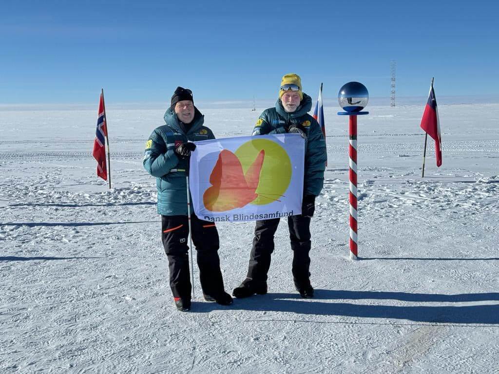 Arne og Claus står på den geografiske Sydpol og holder et flag med Dansk Blindesamfunds logo