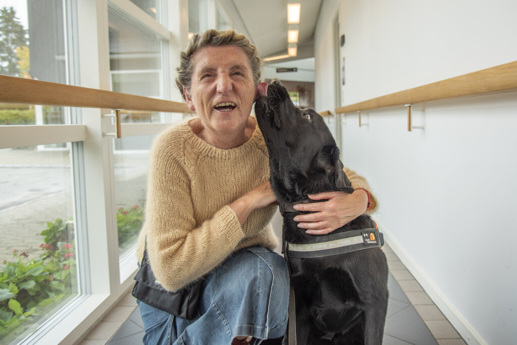 Rita sidder på hug ved siden af sin førerhund Tommy, en sort labrador, der slikker Rita på kinken, som gengælder med et stort grin