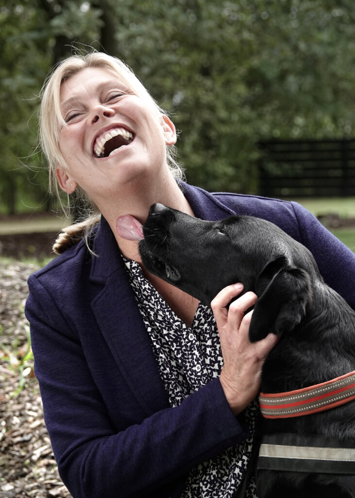 Den sorte labrador Holly giver Foderværtinstruktøren Kirsten et kærligt hundeslik på halsen. Kirsten griner højlydt imens.