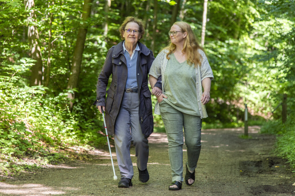 Birgitte Borrebye holder fast i hendes ledsagers arm og har blindestokken i den anden hånd. De går tur i skoven