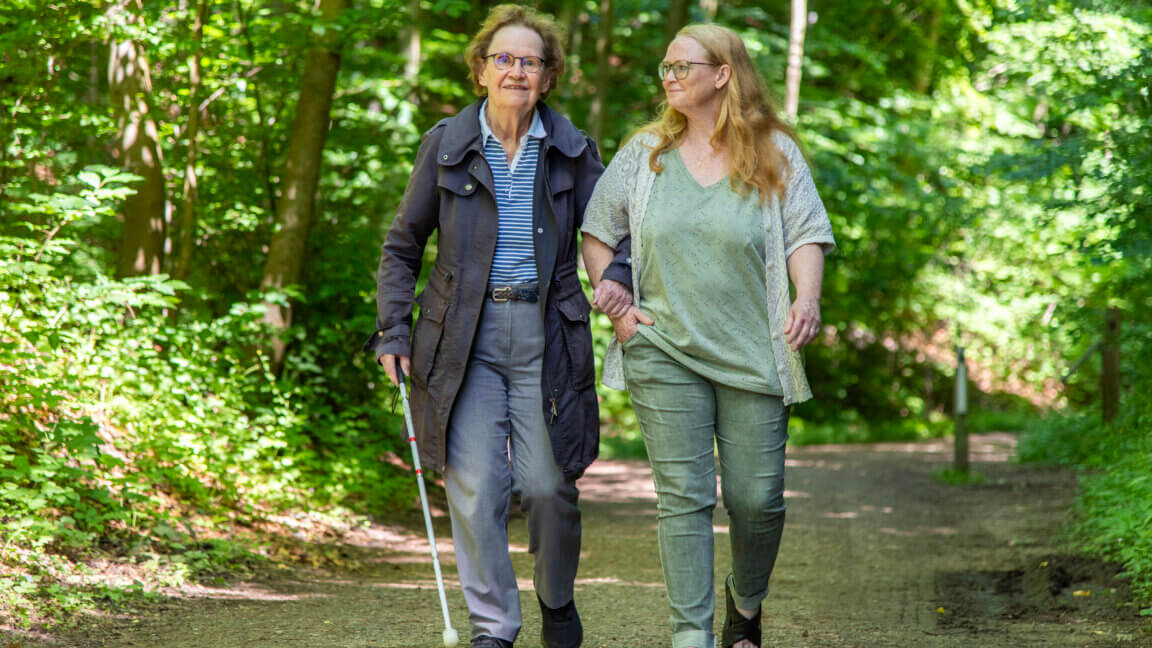 Birgitte Borrebye holder fast i hendes ledsagers arm og har blindestokken i den anden hånd. De går tur i skoven