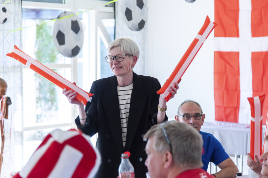 Helle Riley står med to klappepølser, dannebrogsflag på kinderne og i baggrunden hænger guirlande med fodbolde og et stort dannebrog