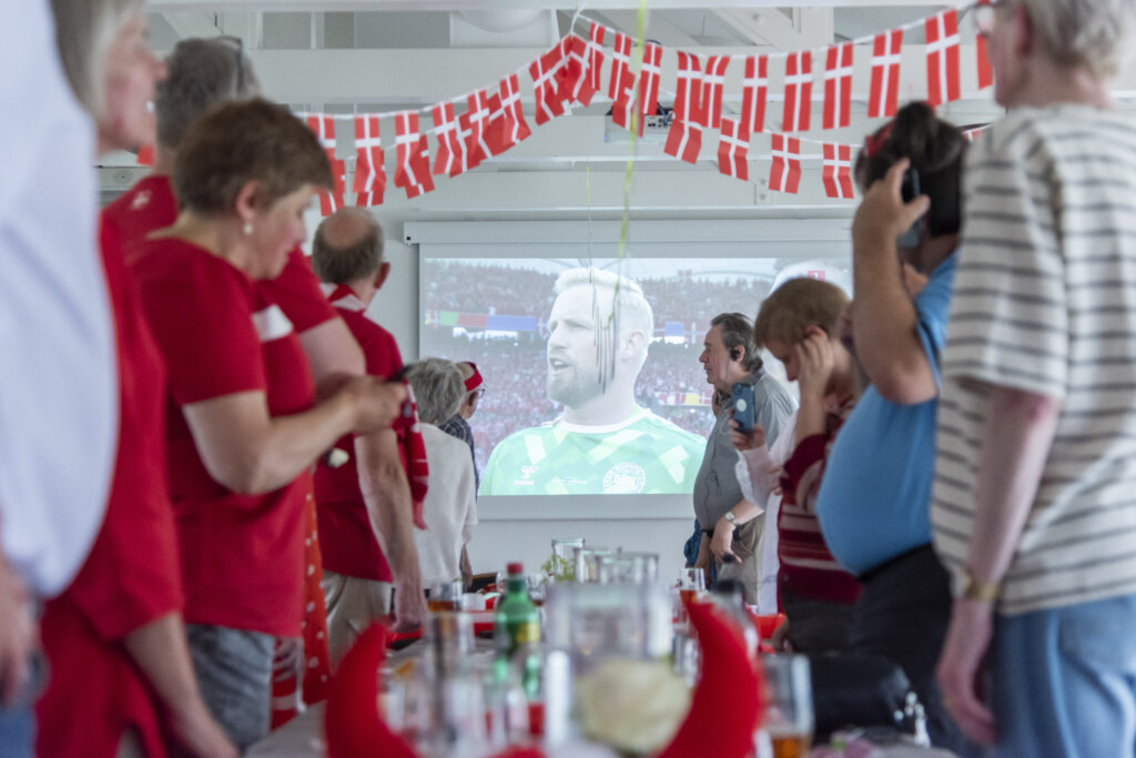 Deltagerne står op til storskærm, der viser fodboldmålmanden Kasper Schmeichel. I loftet hænger guirlande med dannebrogsflag