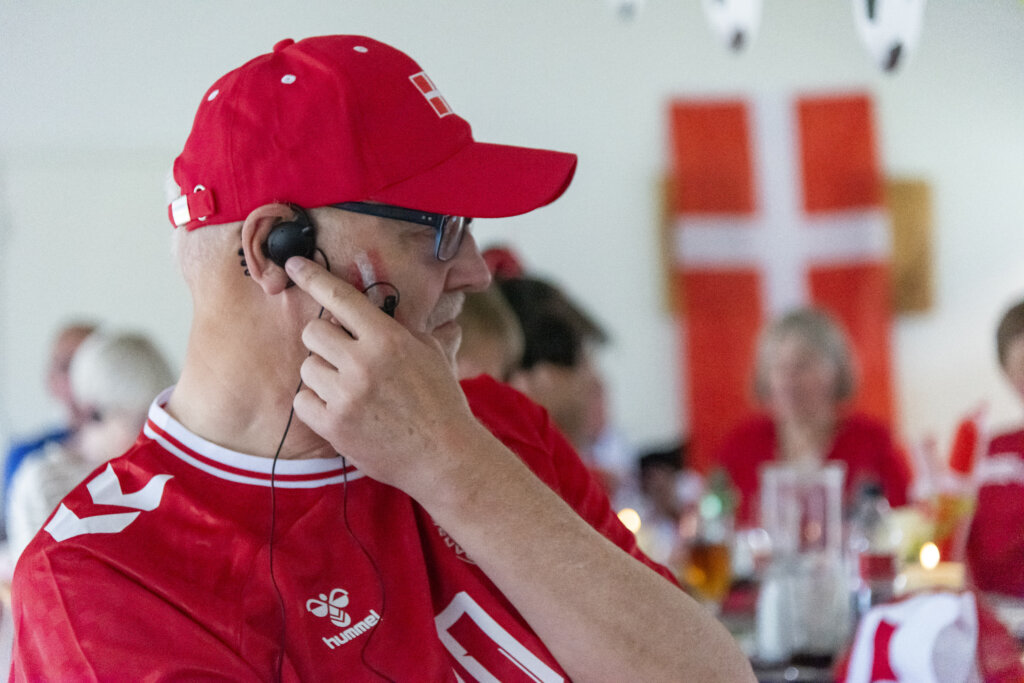 Erik Gottfredsen er iklædt landsholdstrøje, dannebrog på kinderne og rød kasket. Han sætter fingeren til høretelefon i det ene øre