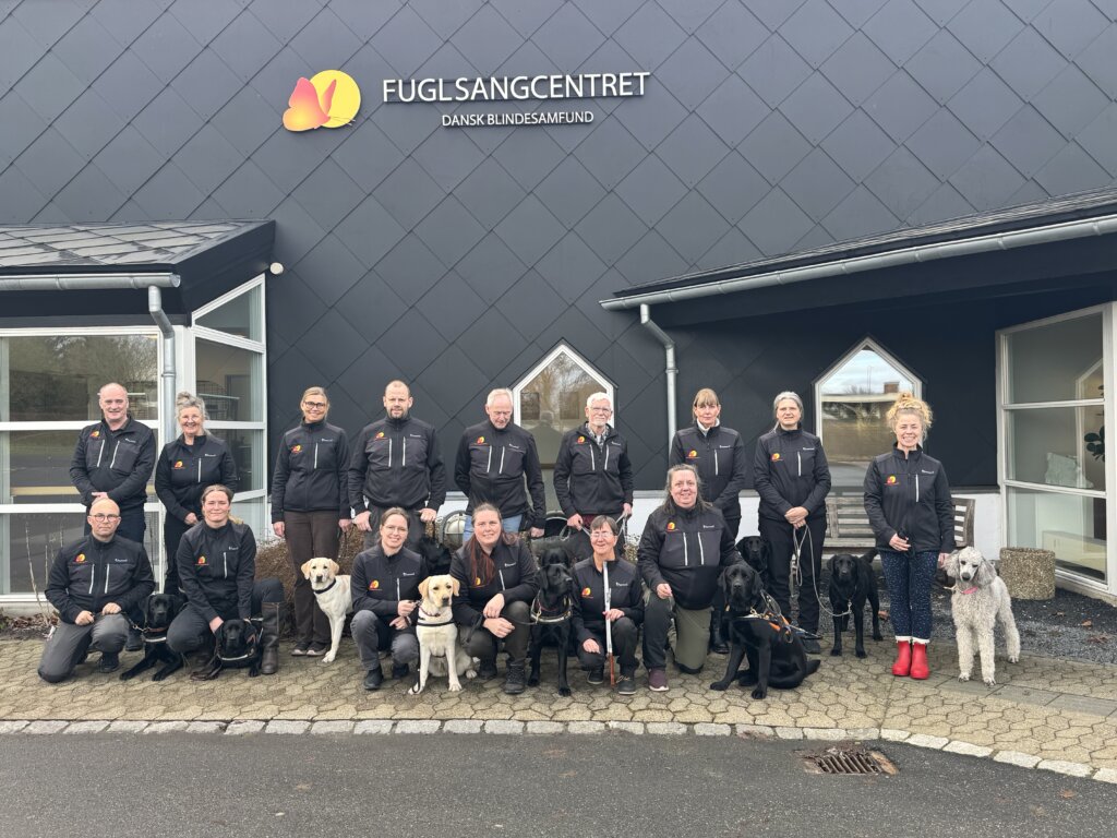 Gruppebillede af førerhundeinstruktører og førerhundekonsulenter i Dansk Blindesamfund. Billedet er taget ude foran Fuglsangcentret. Flere instruktører har førerhunde med på billedet