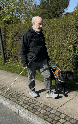 Kurt kommer gående med Jolly, en sort labrador i førerhundebøjle. Kurt med en mobilitystok i den ene hånd og følges pænt med Jolly på fortovet