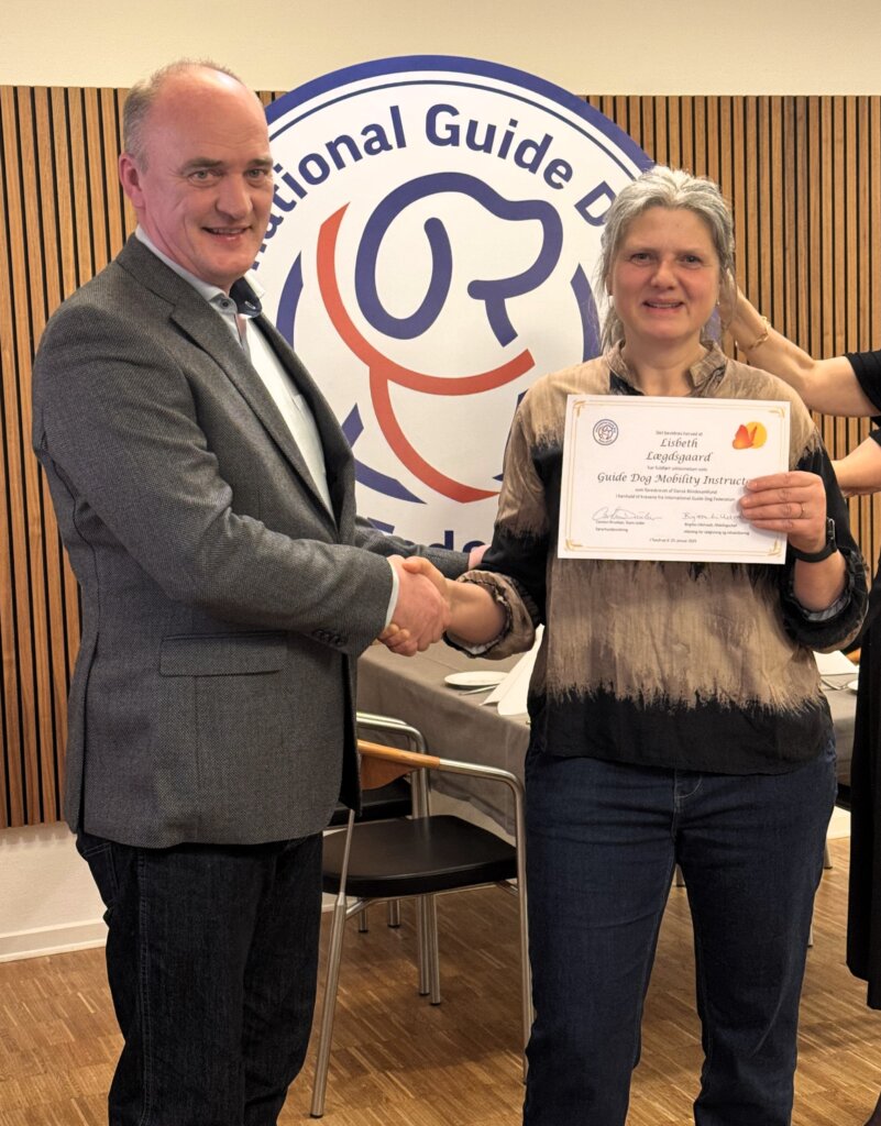 Lisbeth får overrakt diplom på uddannelsen af Carsten Druekær. I baggrunden er et stort skilt med logo fra International Guide Dog Federation