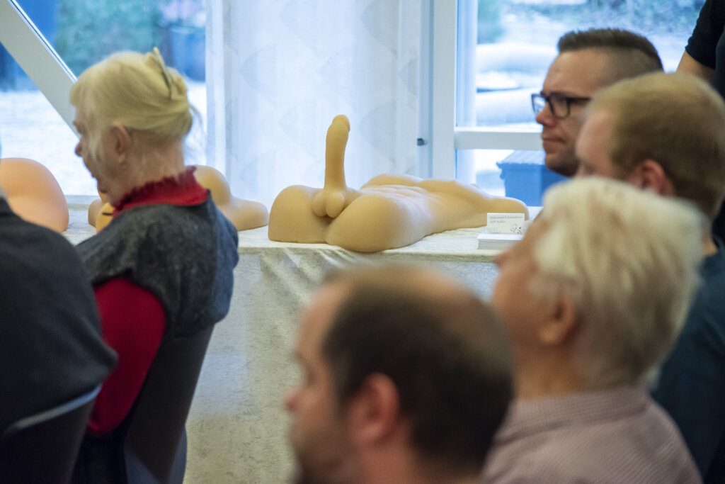 Vi ser deltagere lytte til oplæg. I baggrunden vises en anatomisk model af en penis