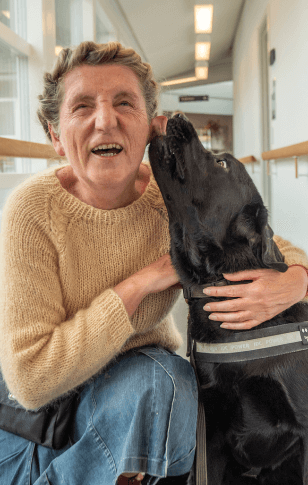 Rita sidder på hug ved siden af sin førerhund Tommy, en sort labrador, der slikker Rita på kinken, som gengælder med et stort grin