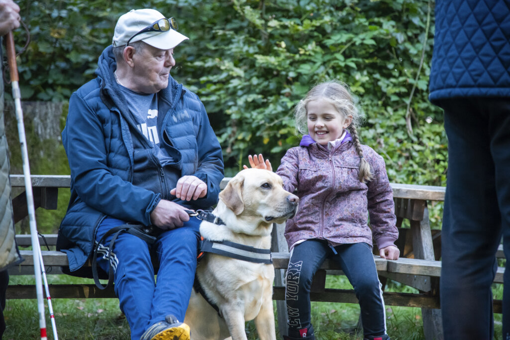 En bedstefar og en pige sidder på en bænk og klapper en førerhund. En lys labrador