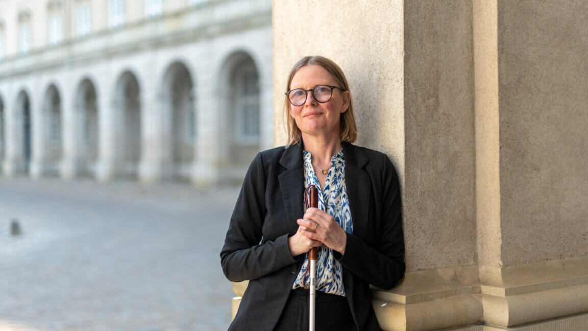 Diana Stentoft står uden for med sin blindestok og læner sig op ad en søjle