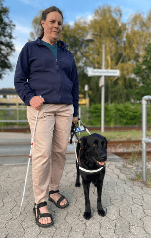 Lene står med førerhunden Hilda, en sort labrador i førerhundebøjle, på en togstation