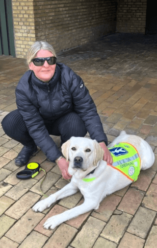 Førerhunden Nova, en lys labrador, ligger på fliser uden for med førerhundevest med Jane ved siden af på hug
