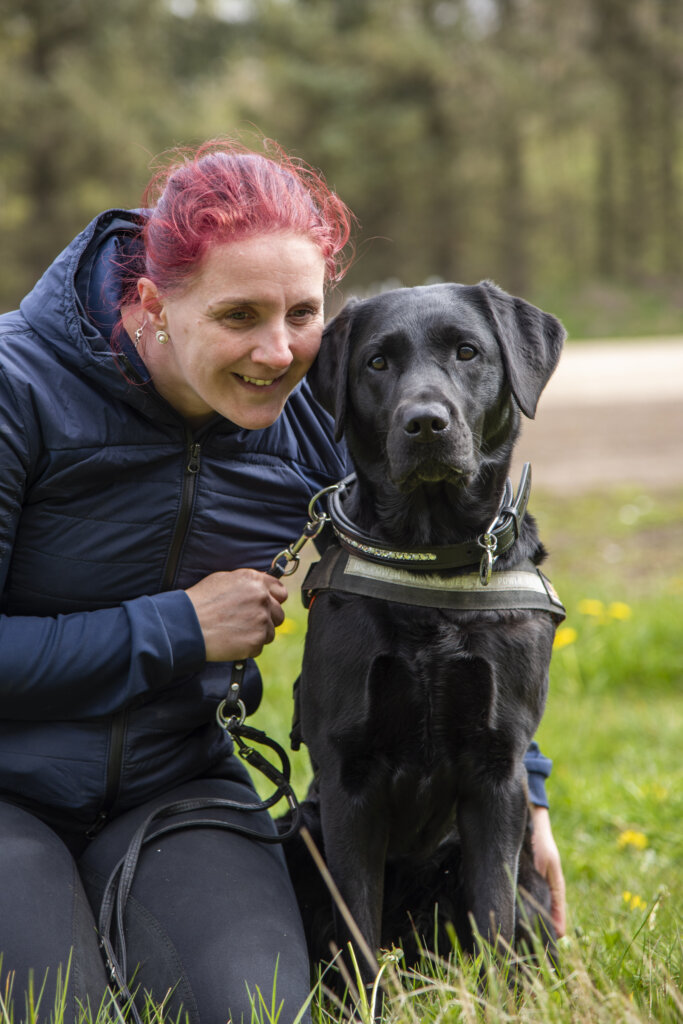 Rikke sidder på hug i græsset uden for og holder om førerhunden Silke, e sort labrador iført førerhundebøjle