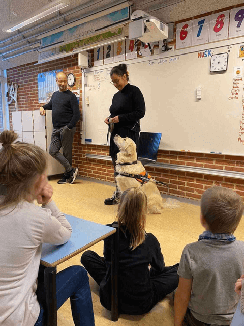 Tijana står foran en skoleklasse med førerhunden Buster ved siden af, en lys golden retriever