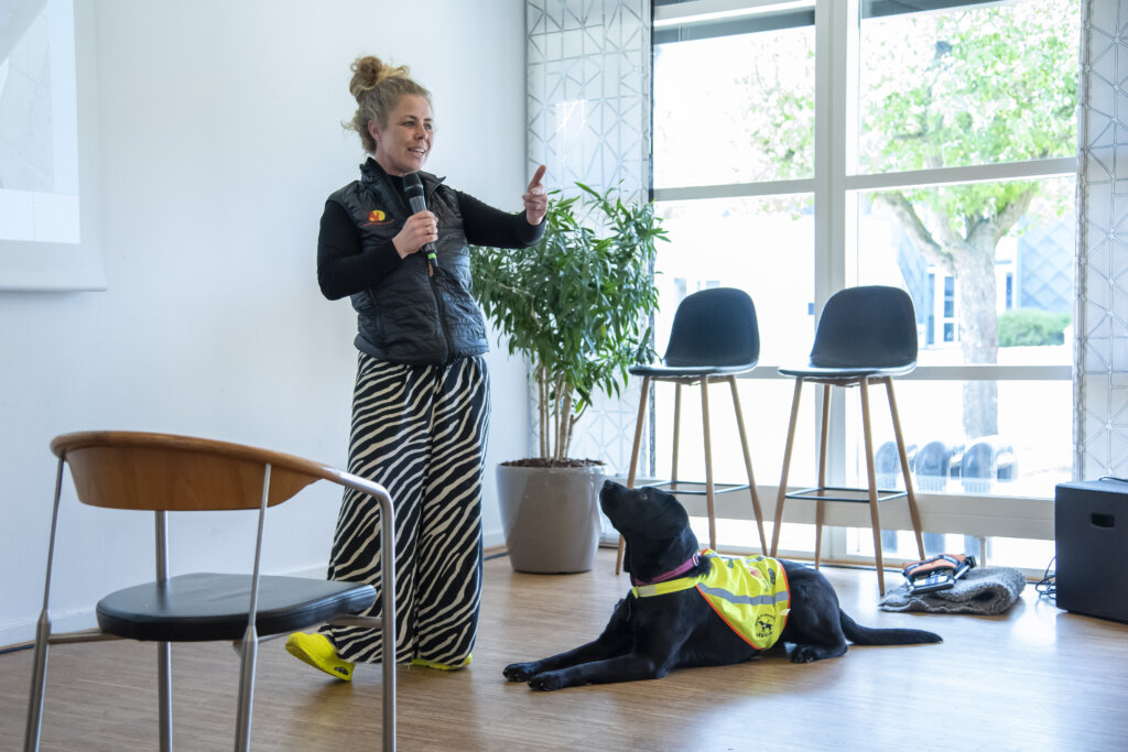 Maria står med en mikrofon og forklarer, hvordan hun træner førerhunde. Ved siden af ligger Vega, en sort labrador med gul førerhundevest, som kigger på hende