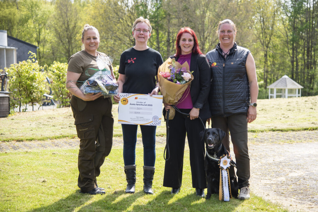 Silkes fodervært, opdrætter og førerhundeinstruktør står ved siden af Rikke og Silke. Rikke holder en buket blomster, opdrætter holder gavekort og fodervært holder gavekurv med godbidder