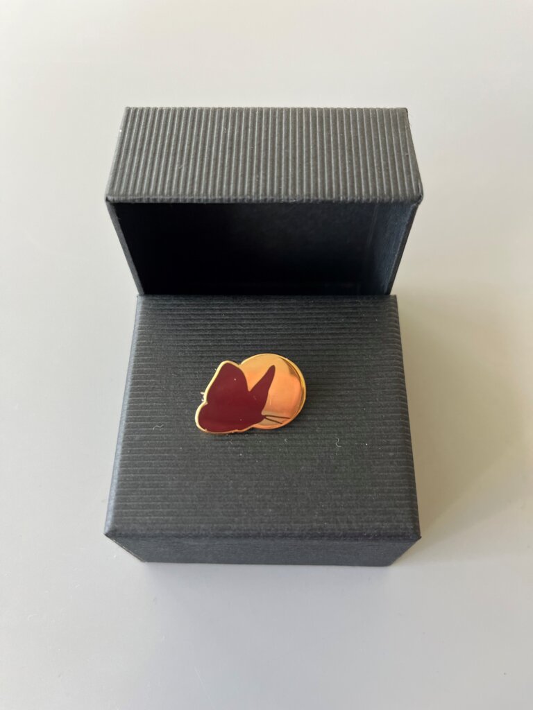 Hæderstegnet i æske. Lavet i guld og forestiller Dansk Blindesamfunds logo med en sommerfugl i orange foran solen i guld