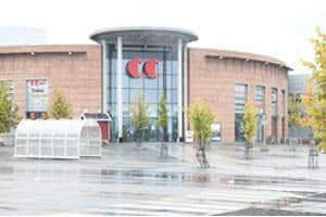 Skarpt billede: Indgang til indkøbscenter med rund glasfacade og murstensbygning, set fra en våd plads foran bygningen.