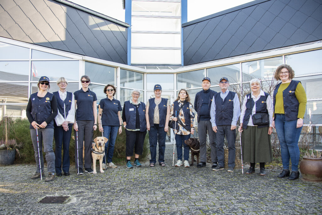 Gruppebillede af Synslinjens frivillige r&aring;dgivere og Helle Riley. De er 11 og to f&oslash;rerhunde, som st&aring;r i en g&aring;rd p&aring; Fuglsangcentret