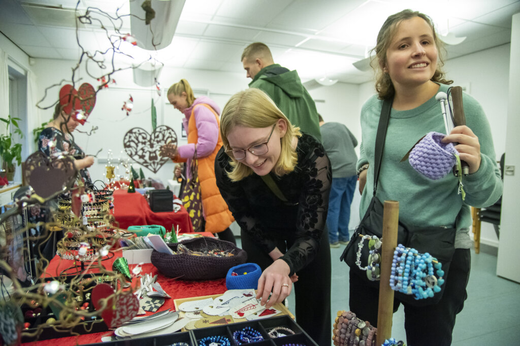 Billedet viser to unge kvinder, der står til et julemarked. De står og kigger på nogle perle armbånd. I Baggrunden ses 4 andre mennesker der går rundt og kigger på julemarkedet.