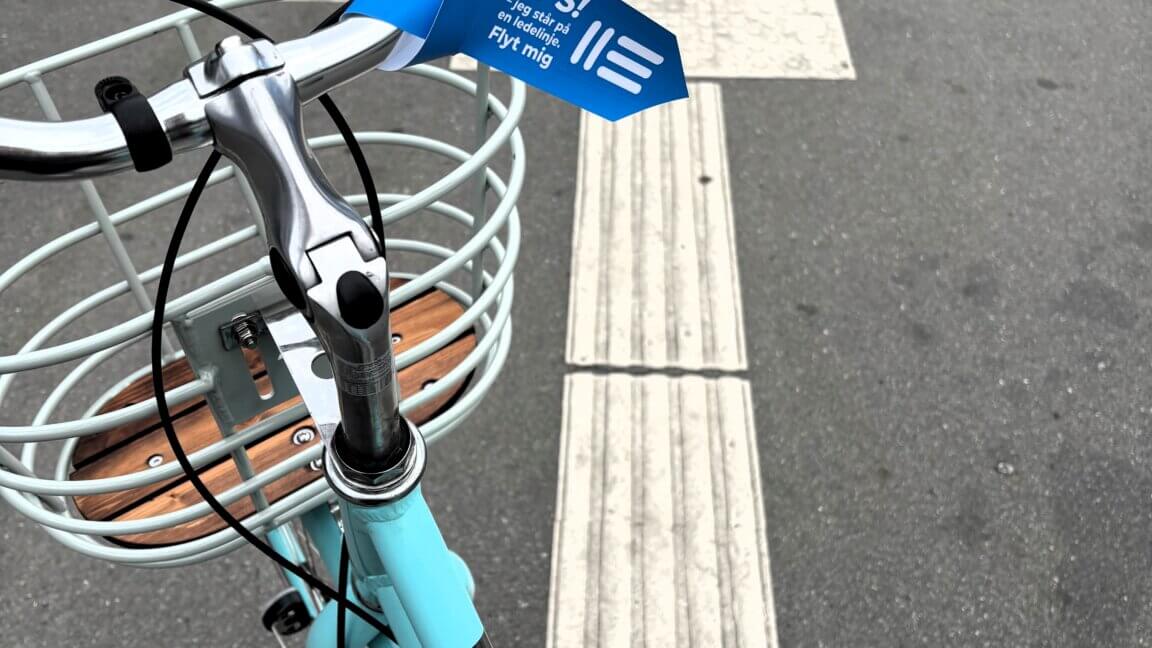 En blå cykel står på tværs af en ledelinje. På styret hænger et skilt, hvor der står: "Ups, jeg står på en ledelinje. Flyt mig"