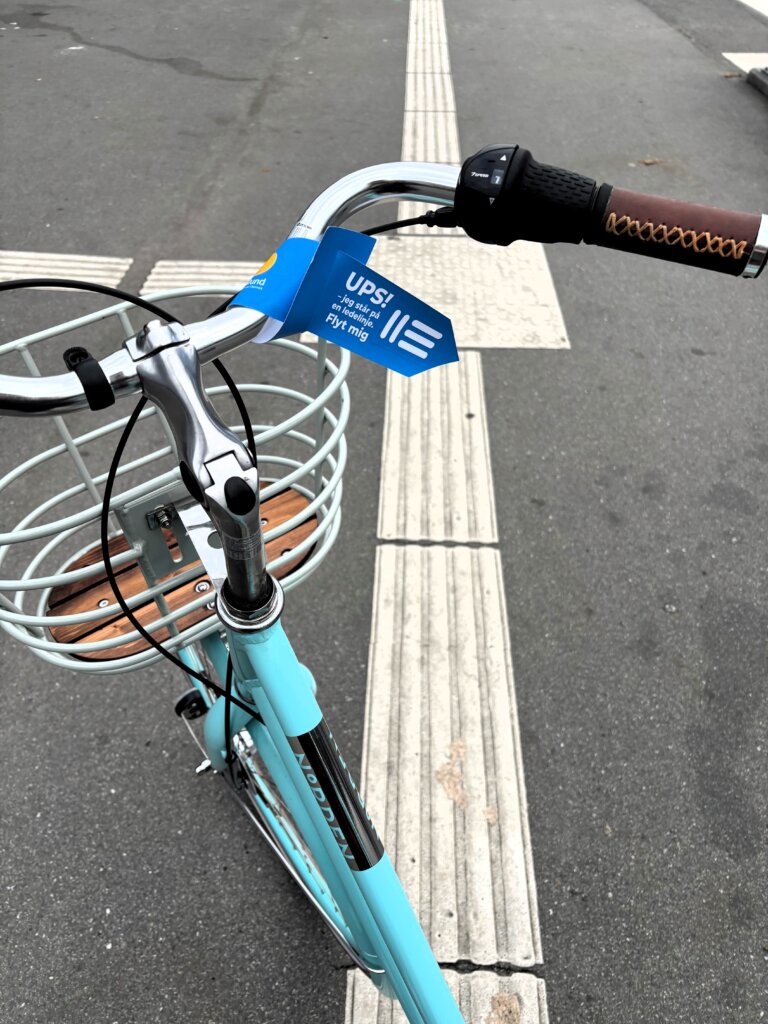 En blå cykel står på tværs af en ledelinje. På styret hænger et skilt, hvor der står: "Ups, jeg står på en ledelinje. Flyt mig"