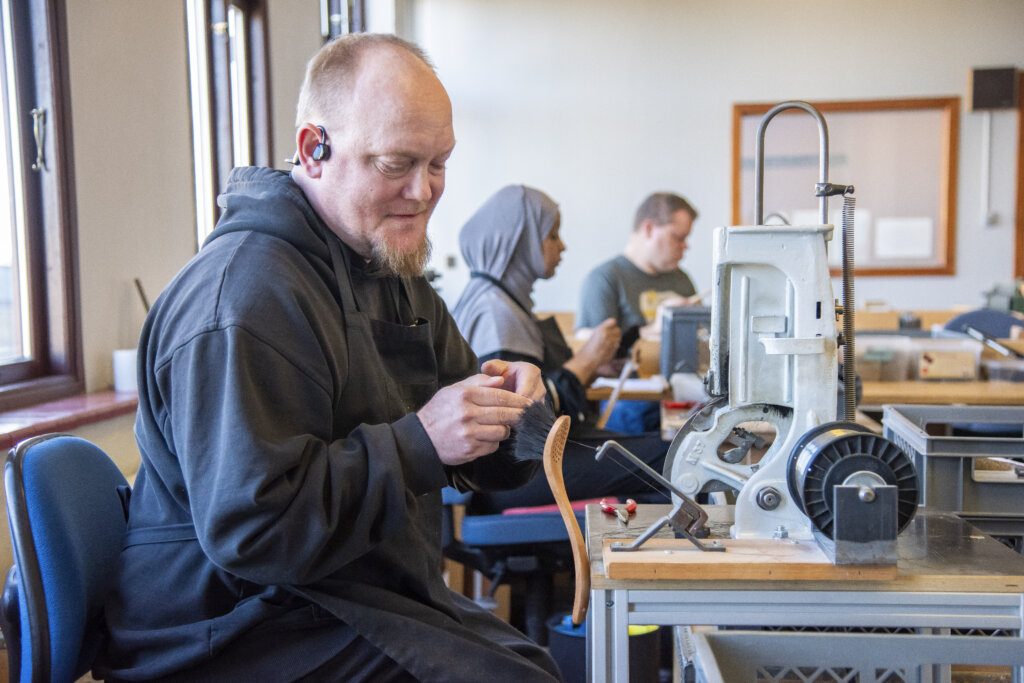Billedet viser Marc, der sidder ned på en stol og er i gang med at lave en opvaskebørste. I baggrunden sidder der en kvinde og en mand der også laver håndarbejde.